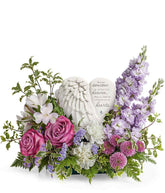 Sympathy flower arrangement with lavender roses, white alstroemeria, white carnations, lavender stock, lavender button spray chrysanthemums, and greenery, surrounding a heart-shaped keepsake with angel wings and an inscription about loved ones in heaven.