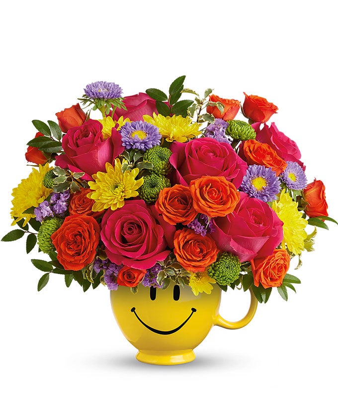 Hot Pink Roses, Orange roses, lavender asters, Yellow & green chrysanthemums, floral greens in a yellow smiley face mug vase against a white background
