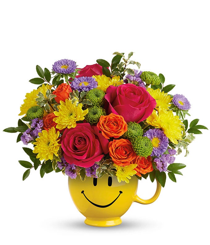 Hot Pink Roses, Orange roses, lavender asters, Yellow & green chrysanthemums, floral greens in a yellow smiley face mug vase against a white background