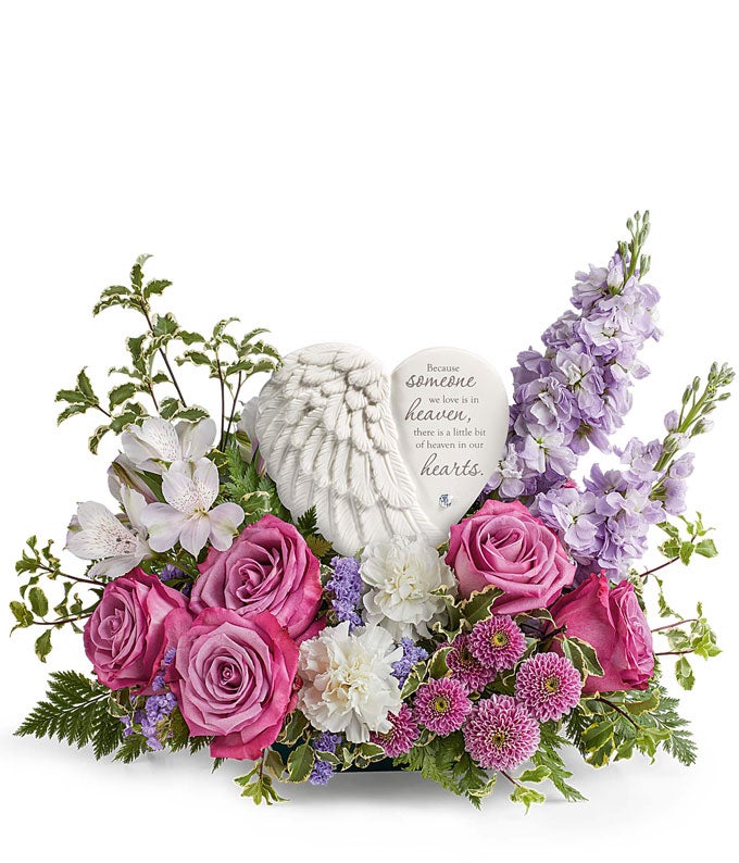 Sympathy flower arrangement with lavender roses, white alstroemeria, white carnations, lavender stock, lavender button spray chrysanthemums, and greenery, surrounding a  heart-shaped keepsake with angel wings and an inscription about loved ones in heaven.