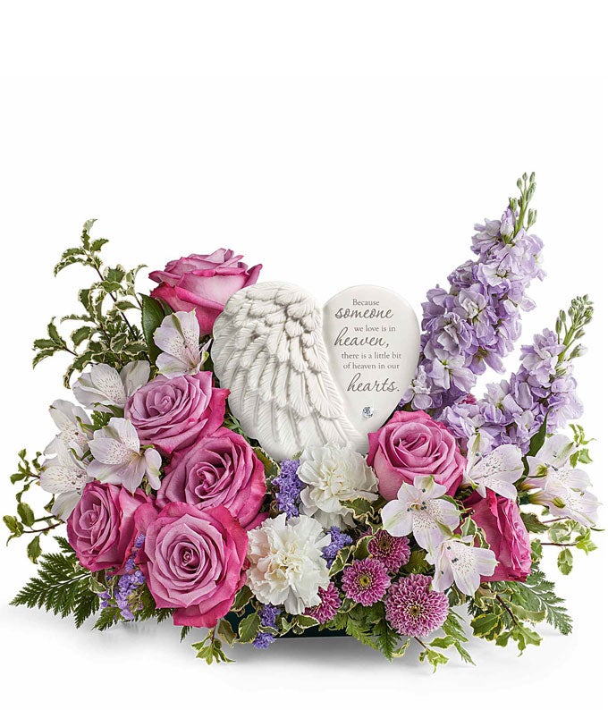 Sympathy flower arrangement with lavender roses, white alstroemeria, white carnations, lavender stock, lavender button spray chrysanthemums, and greenery, surrounding a  heart-shaped keepsake with angel wings and an inscription about loved ones in heaven.