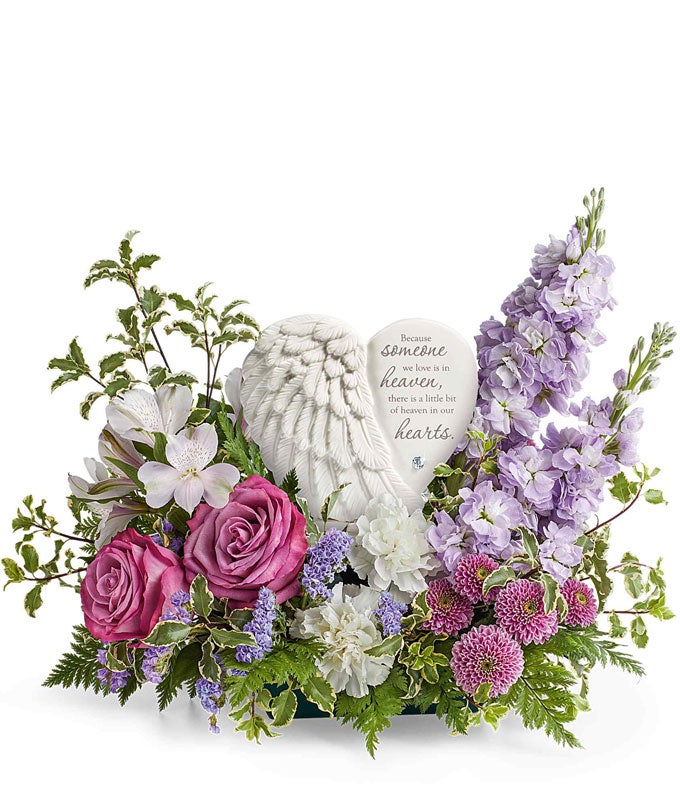 Sympathy flower arrangement with lavender roses, white alstroemeria, white carnations, lavender stock, lavender button spray chrysanthemums, and greenery, surrounding a  heart-shaped keepsake with angel wings and an inscription about loved ones in heaven.