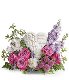 Sympathy flower arrangement with lavender roses, white alstroemeria, white carnations, lavender stock, lavender button spray chrysanthemums, and greenery, surrounding a  heart-shaped keepsake with angel wings and an inscription about loved ones in heaven.